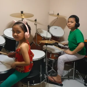Two young students playing drums with headphones on, practicing together and building rhythm skills.