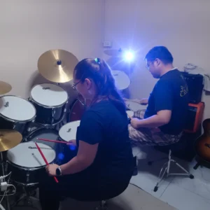 Two students playing drums together in a practice room, showcasing coordination, rhythm, and collaborative music learning.