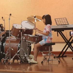 A young girl playing a drum set on stage, practicing percussion skills in a music class or recital setting.